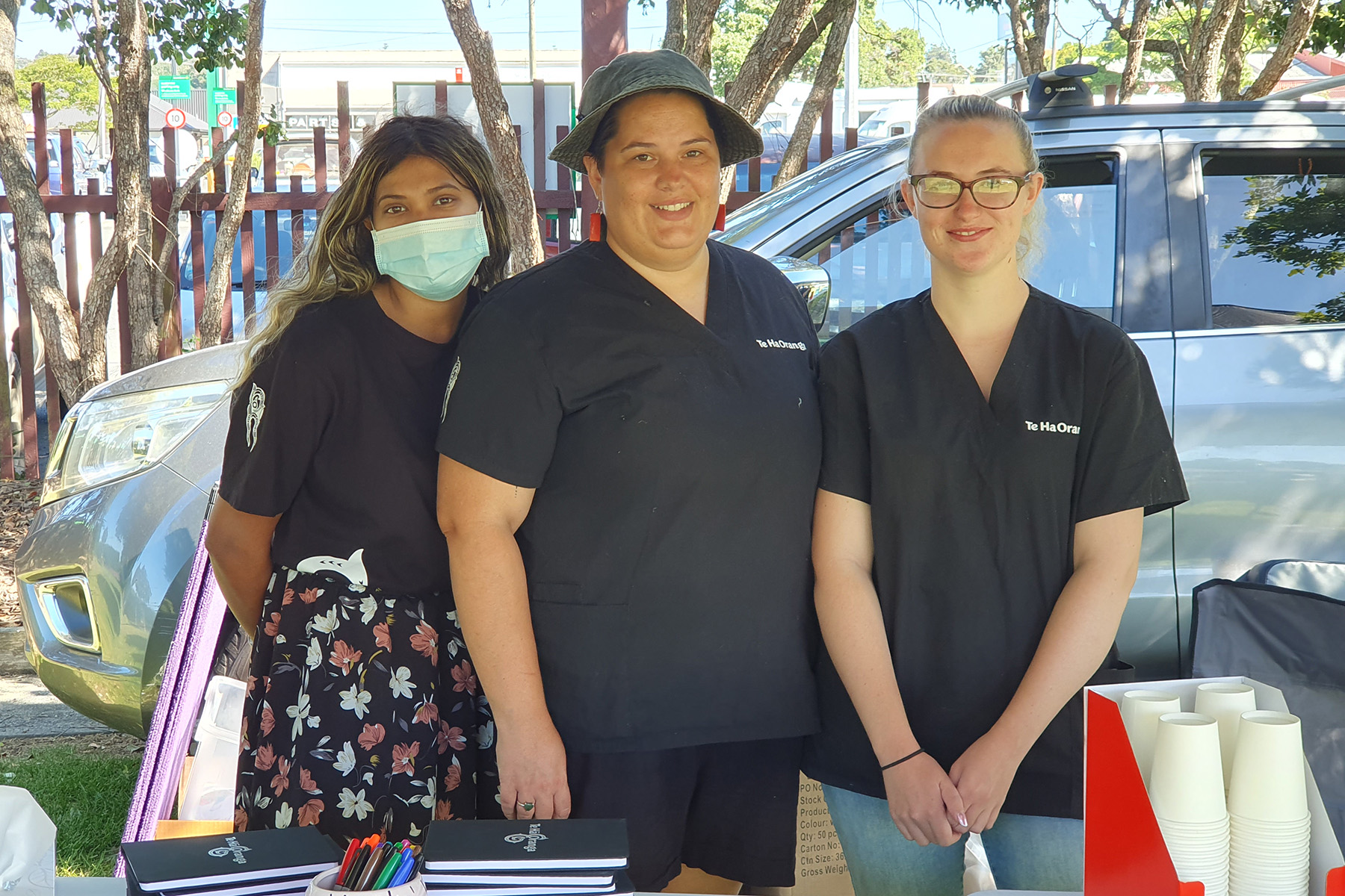 Three adults stand behind a table and smile at the camera. A car, fence and trees are in the background.