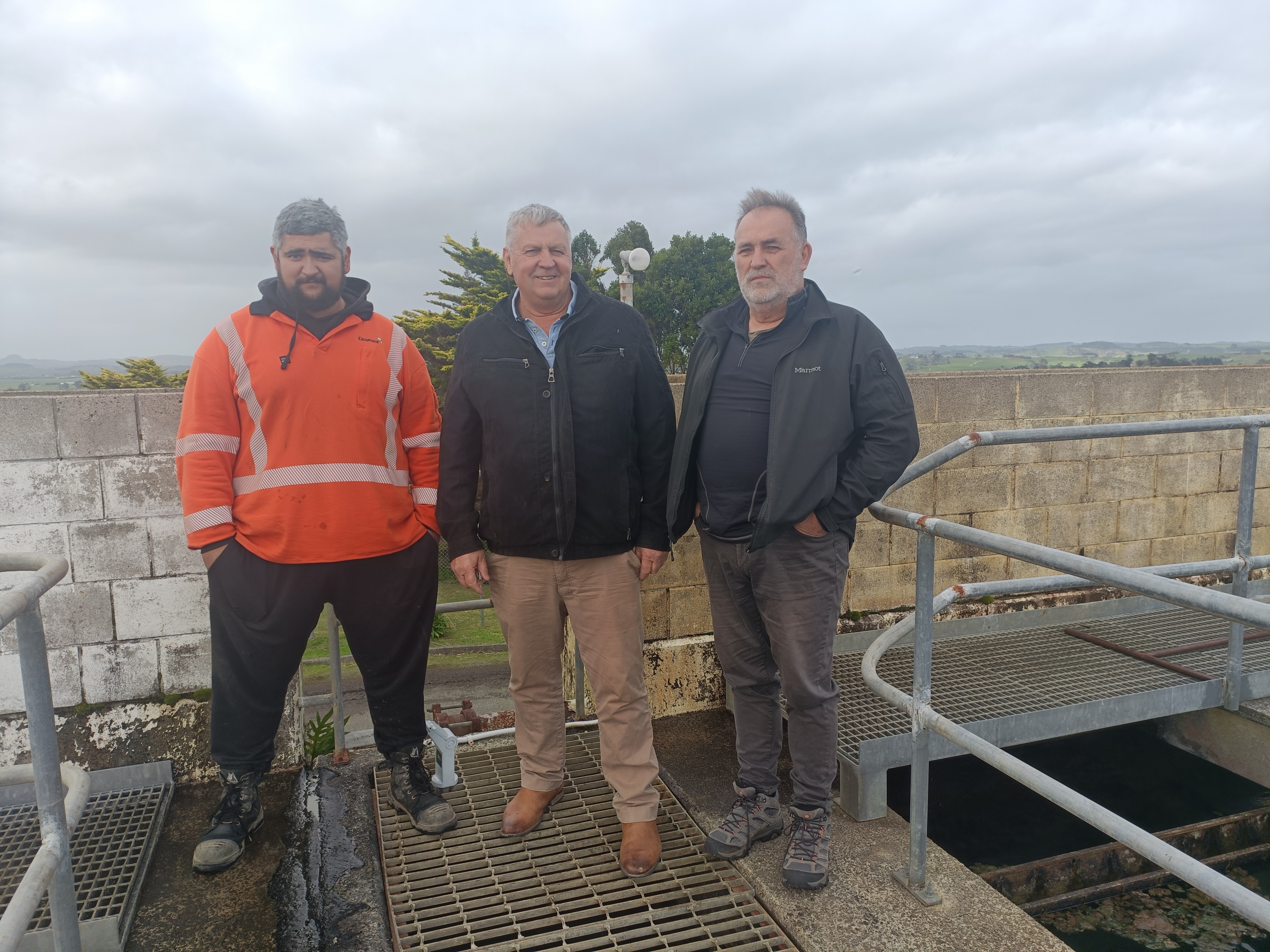 Three men, one in orange hi viz jacket standing on concrete tanks